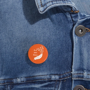 May include: An orange button pin with the text "Every Child Matters" and a white feather and bird graphic. The pin is set against a blue denim jacket.