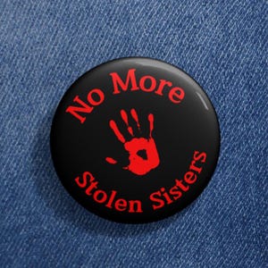 Puede incluir: Un botón negro con el texto "No More Stolen Sisters" en letras rojas. Una huella de mano roja está en el centro del botón.