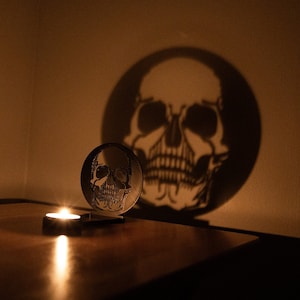 May include: A black metal skull candle holder casts a shadow of a skull on the wall. A lit candle sits in the holder on a wooden table.