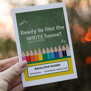 May include: A white rectangular card with a green chalkboard background. The card has the text "Ready to find the WRITE home?" and "100%" in white. A row of colorful pencils is at the bottom, with a yellow and green section that says "REALTOR NAME".