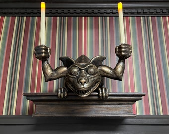 Puede incluir: Candelabro de gárgola de color bronce con dos velas falsas. La gárgola tiene una expresión amenazante y sostiene las velas en alto. El candelabro está montado en un estante de madera oscura, sobre un fondo de papel pintado a rayas.