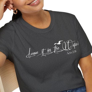 Op de afbeelding: Een donker grijs t-shirt met een witte afbeelding met de tekst "Leave it in the water!" en "Acts 2:18".
