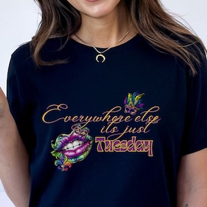 May include: Black t-shirt with the text "Everywhere else it's just Tuesday" in gold and purple script. The shirt features a colorful lip graphic and a butterfly design. The model is wearing a gold necklace and blue jeans.
