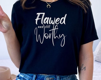 May include: A black t-shirt with white text that reads "Flawed and still worthy".