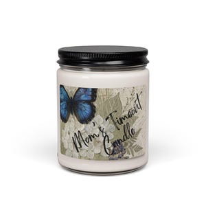 May include: A white candle jar with a black lid. The label features a blue butterfly, white flowers, and the text "Mom's Timeout Candle" in a script font. The candle is a decorative item.