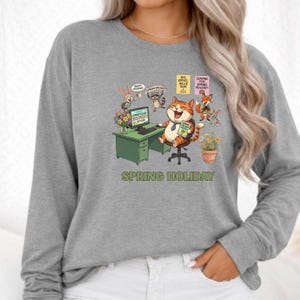 May include: Gray sweatshirt with a colorful graphic featuring a cartoon cat at a desk, surrounded by other cartoon animals. The desk has a computer with the word "BOOKED" on the screen. The text "SPRING HOLIDAY" is printed below the graphic.