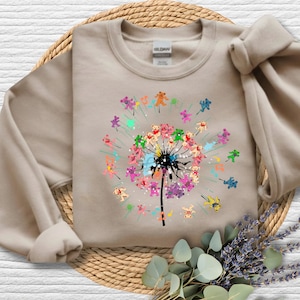 May include: Beige sweatshirt featuring a colorful dandelion design. The dandelion's head is composed of teddy bears in various colors, with musical notes and colorful accents. The sweatshirt has long sleeves and a crew neck.
