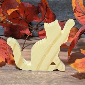 May include: A wooden cat figurine in a playful pose, with its paw raised. The light-colored wood has visible grain and is layered. The cat is set against a backdrop of fall foliage with red and orange leaves.