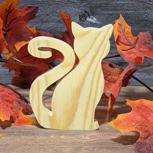 May include: A wooden cat silhouette with a curled tail, crafted from light-colored wood. The cat is surrounded by colorful autumn leaves in shades of red, orange, and brown, creating a seasonal theme. The cat is a decorative item.