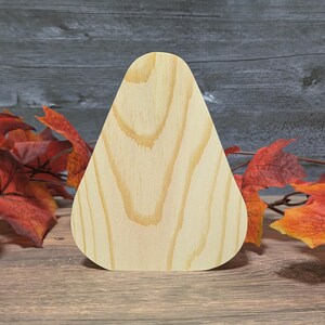 May include: A light-colored, teardrop-shaped wooden cutout with visible wood grain. The cutout is displayed on a wooden surface, with fall-colored leaves in the background. The cutout is likely for crafting or decorative purposes.