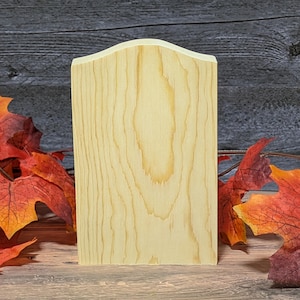 May include: A rectangular piece of light-colored, unfinished wood with a curved top edge. The wood displays a natural grain pattern with vertical lines running throughout the surface, showcasing the texture and light tan color of the material.