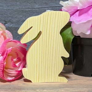 May include: A wooden bunny figurine with a light yellow finish. The bunny is standing upright with its ears perked up. The figurine is next to pink and white flowers in a black pot. The wood grain is visible.
