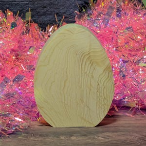 May include: A light-colored, wooden egg-shaped object with visible wood grain. The egg is standing upright against a backdrop of pink and iridescent tinsel. The wood has a natural, unfinished appearance.