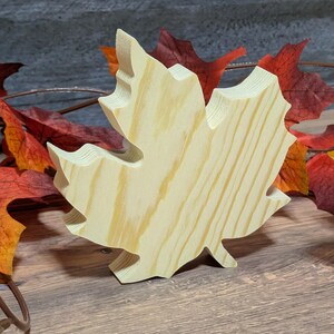 May include: A wooden maple leaf cutout, displaying natural wood grain and a three-dimensional design. The leaf is light-coloured, with visible wood grain, and is surrounded by colourful autumn leaves, creating a seasonal theme.