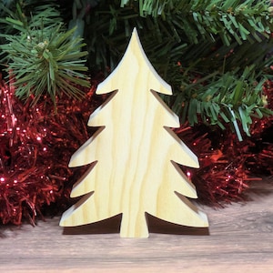 May include: A wooden Christmas tree ornament with a natural wood grain finish. The tree is cut in a classic evergreen shape, with layered branches. The background features Christmas decorations, including red tinsel and green pine branches.