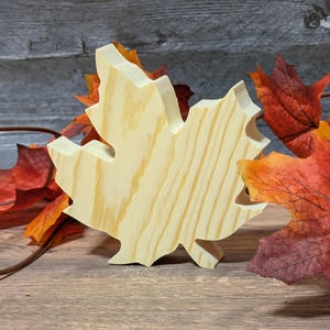 May include: A wooden maple leaf cutout with visible wood grain, standing upright. The leaf is light-coloured and surrounded by artificial autumn leaves in shades of red, orange, and yellow, suggesting an autumn theme.