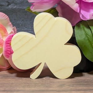 May include: A wooden shamrock cutout with a light, natural wood finish. The three-leaf clover shape is displayed against a backdrop of pink flowers and green leaves, suggesting a St. Patrick's Day theme or general nature decor.