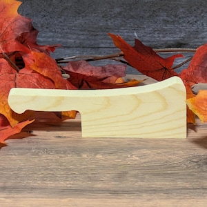 May include: A light-colored wooden cutting board with a handle. The board is rectangular with a curved handle on the left side. The background features fall leaves in shades of red and orange.