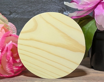 Puede incluir: Un disco de madera redondo sin terminar con vetas visibles. El disco es de color amarillo claro con vetas marrones. Está rodeado de flores rosas y blancas y una maceta negra.