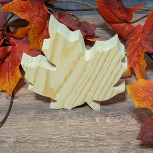 May include: A wooden maple leaf decoration with a light wood grain finish. The leaf is surrounded by autumn-coloured artificial leaves in shades of red, orange, and brown. The leaf is standing on a wooden surface.