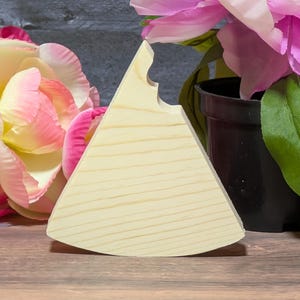 May include: A light-colored wooden wedge with a notched edge, possibly a cheese board or serving piece. The wood grain is visible. The wedge is positioned in front of pink and yellow flowers and a black pot.