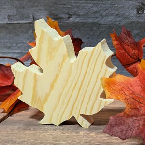 May include: A wooden maple leaf cutout with visible wood grain. The leaf is light-coloured and stands upright. Artificial autumn leaves in shades of red, orange, and yellow are in the background, suggesting an autumn theme.