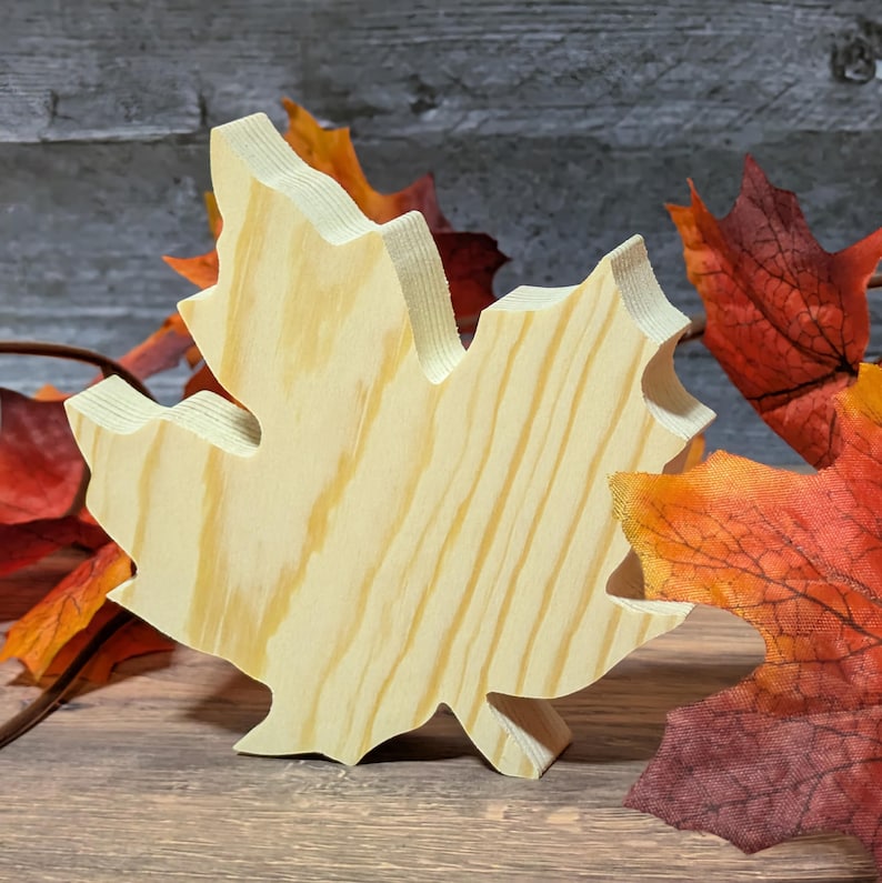 May include: A wooden maple leaf cutout with visible wood grain. The leaf is light-coloured and stands upright. Artificial autumn leaves in shades of red, orange, and yellow are in the background, suggesting an autumn theme.