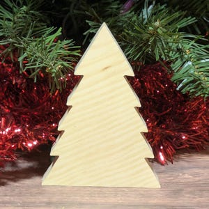 May include: A wooden Christmas tree decoration. The tree is cut from light-colored wood and has a simple, geometric design. The tree is standing on a wooden surface, with red tinsel and green garland in the background.