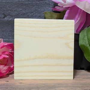 May include: A square, light-colored wooden block with visible wood grain. The block is positioned in front of a blurred background featuring pink and white flowers and green leaves. The wood has a natural, unfinished appearance.