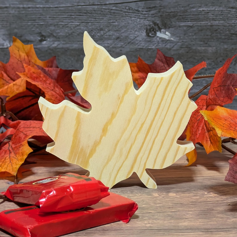 May include: A wooden maple leaf cutout with visible wood grain, positioned in front of autumnal artificial leaves. Two red wrapped packages are in the foreground, suggesting a seasonal theme.