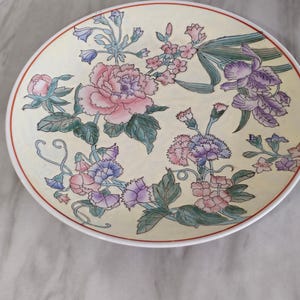 Puede incluir: Un plato decorativo redondo de color crema con un borde rojo. El plato está adornado con un diseño floral con flores rosas, moradas y azules y hojas verdes. Las flores están dispuestas sobre la superficie del plato.