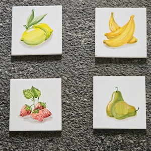 May include: Four white ceramic tiles with hand-painted fruit illustrations. The top left tile features two yellow lemons with green leaves. The top right tile shows three yellow bananas. The bottom left tile has three red strawberries with green leaves. The bottom right tile shows two green pears.