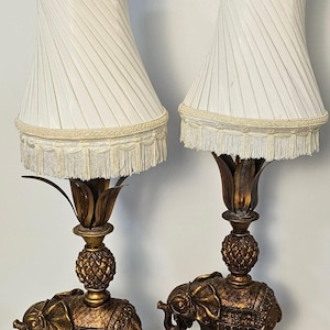 Puede incluir: Par de lámparas de mesa vintage con bases doradas en forma de elefante y pantallas plisadas blancas. Las lámparas tienen un detalle en forma de piña y una franja decorativa. Cada lámpara tiene un remate marrón.