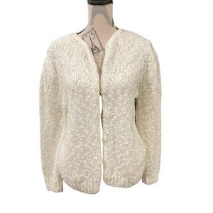 Puede incluir: Cárdigan crema con frente abierto. El suéter tiene un patrón de punto texturizado y mangas largas. Se abotona en la parte delantera y se exhibe en un maniquí. El suéter es de color neutro y adecuado para usar en capas.