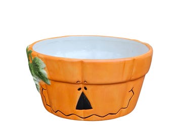 May include: A ceramic pumpkin-shaped bowl with a white interior. The orange exterior features a carved jack-o'-lantern face with a black triangle nose and a jagged mouth. Green leaves and vines are on the side. A festive Halloween decoration.