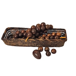 May include: A rectangular woven dark brown rattan basket containing a cluster of smooth, spherical dark brown wooden grapes. Several individual wooden spheres and one larger oval-shaped wooden piece are positioned in front of the basket.
