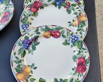 May include: Set of three white ceramic dinner plates with a colorful fruit and floral design. The plates feature a border of green leaves, red apples, purple grapes, and blue plums.