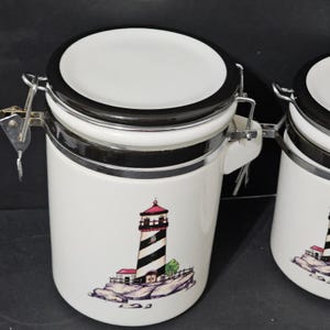 Puede incluir: Bote de cerámica blanco con un diseño de faro en blanco y negro. El bote tiene una tapa blanca con borde negro y un cierre de metal plateado. El faro es blanco y negro con la parte superior roja y está sobre una base rocosa.