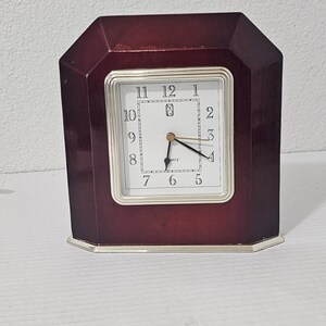 May include: A dark red wooden clock with a silver trim and a white face. The clock has black hands and black Roman numeral markings. The clock face also has the word "ANTZ" printed on it.