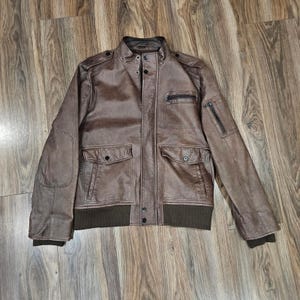 May include: A brown leather jacket with a bomber-style design. The jacket features a stand-up collar, multiple pockets, and a ribbed hem and cuffs. The jacket has a zipper closure and button details.
