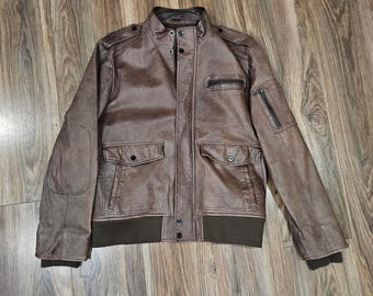 Könnte beinhalten: Braune Lederjacke im Bomber-Stil. Sie hat einen Stehkragen, mehrere Taschen und gerippte Säume. Die Jacke hat einen Reißverschluss und Knöpfe. Ein klassisches Kleidungsstück für viele Gelegenheiten.