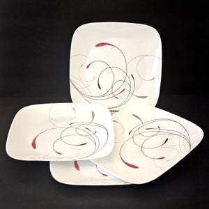 Może przedstawiać: Zestaw czterech kwadratowych białych talerzy ceramicznych z dekoracyjnym wzorem cienkich, płynnych czarnych i ciemnoczerwonych linii, które zakrzywiają się na powierzchni. Talerze mają zaokrąglone rogi oraz gładkie, błyszczące wykończenie.
