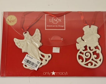 May include: Two ivory-coloured Lenox Christmas ornaments: an angel and a snowman, presented on a red card. The angel has outstretched wings and a flowing gown. The snowman wears a hat and scarf. Both have red ribbon hangers.