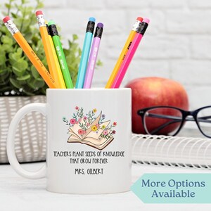 May include: White ceramic coffee mug with a floral design and an open book with the text "Teachers plant seeds of knowledge that grow forever. Mrs. Gilbert."