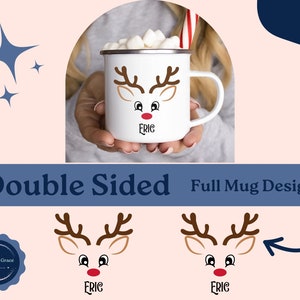 May include: White ceramic mug with a reindeer face design. The reindeer has brown antlers, brown eyes, and a red nose. The mug has the name "Eric" written below the reindeer face. The mug is shown being held by a person's hand.