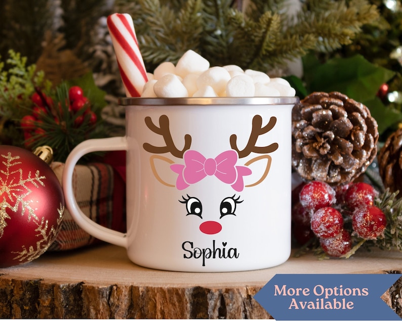May include: White enamel mug with a reindeer face design, a pink bow, and the name "Sophia" printed on the bottom. The mug is filled with marshmallows and a red and white candy cane.