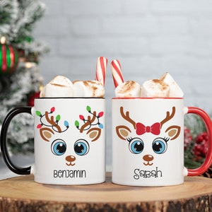 May include: Two white ceramic mugs with red and black handles. The mugs feature a cartoon reindeer design with Christmas lights and the names "Benjamin" and "Sarah" printed on the front.