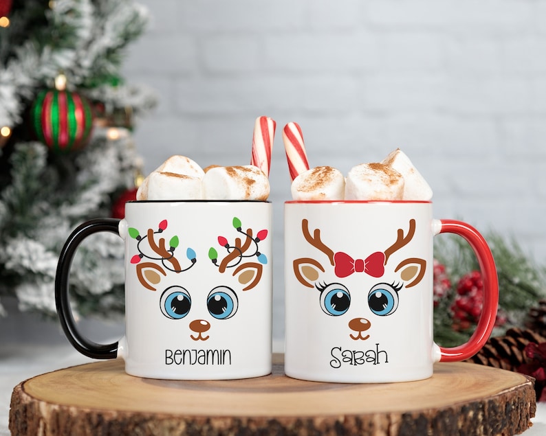May include: Two white ceramic mugs with red and black handles. The mugs feature a cartoon reindeer design with Christmas lights and the names "Benjamin" and "Sarah" printed on the front.