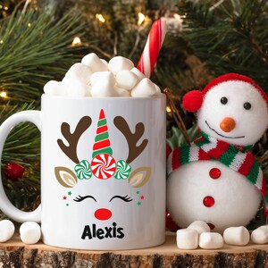 May include: White ceramic mug with a reindeer unicorn design, red nose, green and red candy cane antlers, and the name "Alexis" printed on the bottom. The mug is filled with marshmallows and a candy cane.