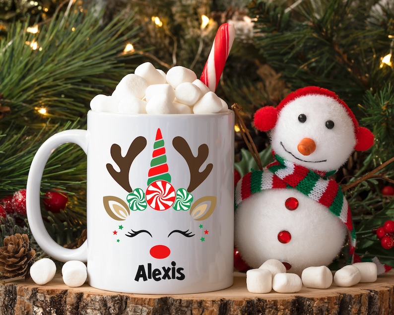 May include: White ceramic mug with a reindeer unicorn design, red nose, green and red candy cane antlers, and the name "Alexis" printed on the bottom. The mug is filled with marshmallows and a candy cane.